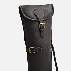 Può includere: Una custodia per fucile in pelle nera con chiusura a patta e fibbie in ottone. La custodia ha una tracolla e una cerniera laterale. La pelle ha un aspetto strutturato.