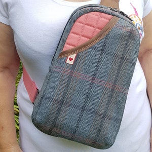 May include: A grey and pink crossbody bag with a quilted pink pocket and a brown leather trim. The bag has a pink adjustable strap and a small red heart patch.