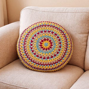 May include: A round, crocheted pillow with a colorful mandala design. The pillow features concentric circles of red, yellow, blue, and tan yarn. The pillow is placed on a beige sofa, creating a cozy and decorative accent.