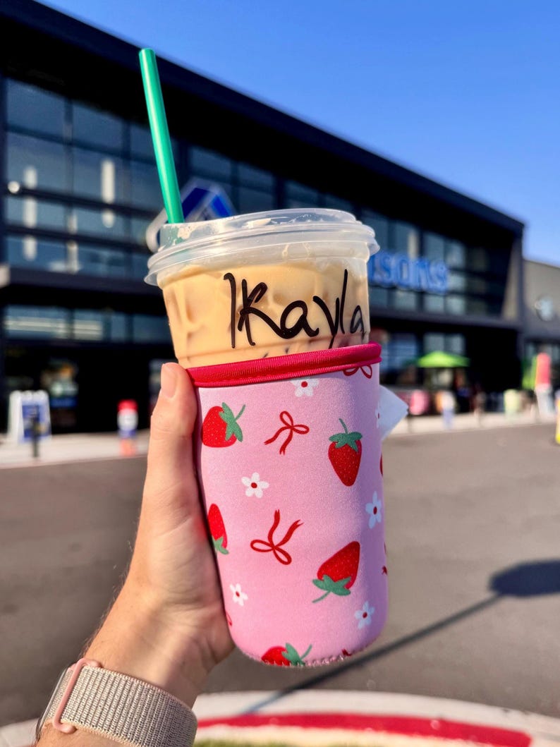 Pu&ograve; includere: Un bicchiere di plastica trasparente con caff&egrave; freddo e una cannuccia verde, tenuto in una custodia rosa a forma di fragola. Il nome "Kayla" &egrave; scritto sul bicchiere. La custodia presenta fragole rosse, fiocchi e fiori bianchi.