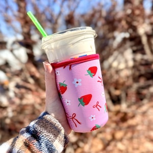 Pu&ograve; includere: Una custodia rosa per bevande con un motivo a fragole, che tiene in mano un bicchiere di plastica trasparente con una bevanda bianca e una cannuccia verde. La custodia ha bordi rossi ed &egrave; decorata con fragole rosse, fiori bianchi e fiocchi rossi.