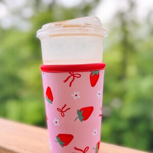 Pu&ograve; includere: Un bicchiere di plastica trasparente con coperchio &egrave; all'interno di una custodia rosa. La custodia &egrave; decorata con fragole rosse, fiori bianchi e fiocchi rossi. La parte superiore della custodia ha un bordo rosso.