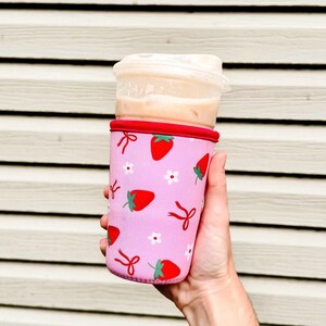 Pu&ograve; includere: Una custodia rosa per bevande con finiture rosse, ornata da un motivo di fragole rosse, fiori bianchi e fiocchi rossi. La custodia contiene un bicchiere di plastica trasparente con una bevanda all'interno.