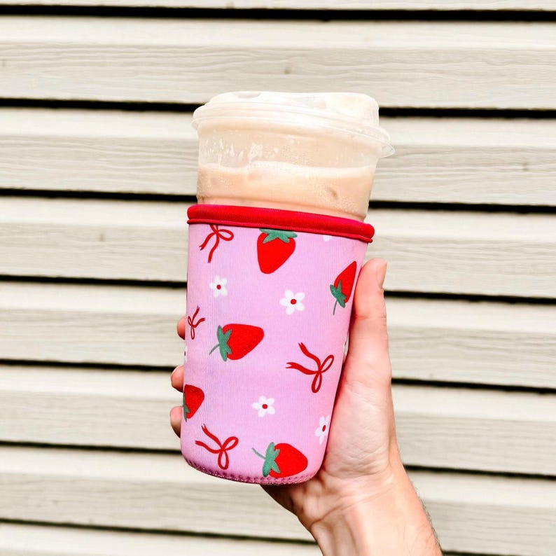 Pu&ograve; includere: Una custodia rosa per bevande con finiture rosse, ornata da un motivo di fragole rosse, fiori bianchi e fiocchi rossi. La custodia contiene un bicchiere di plastica trasparente con una bevanda all'interno.