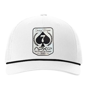 Pode incluir: Boné branco de caminhoneiro com aba preta e detalhe de cordão preto. A frente apresenta um patch com o texto "Sonny Hayes" acima de um espadas preto com o número 7, e o texto "APX GP CHAMP - 2023".