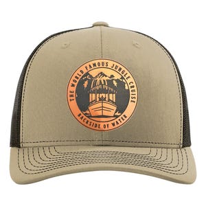 Könnte beinhalten: Beige Trucker-Cap mit schwarzem Mesh-Rücken und gebogenem Schirm. Auf der Vorderseite befindet sich eine runde Grafik mit dem Text "THE WORLD FAMOUS JUNGLE CRUISE BACKSIDE OF WATER" und einer Bootsillustration in Orange und Schwarz.