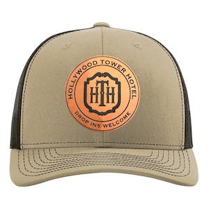 May include: A tan and black trucker hat with a circular patch featuring the text "HOLLYWOOD TOWER HOTEL" and "DROP INS WELCOME". The patch has a copper-colored background with a stylized "HTH" logo.