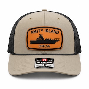 May include: A beige and black trucker hat featuring a rectangular orange patch on the front. The patch displays a black silhouette of a boat with the text AMITY ISLAND and ORCA. A black sticker on the brim reads RICHARDSON 112 TRUCKER.