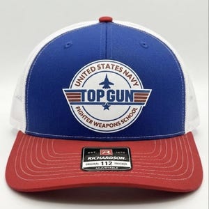 May include: A blue, white, and red Top Gun trucker cap. The front panel is blue with a white circular emblem that reads "United States Navy Top Gun Fighter Weapons School." The peak is red, and the mesh back is white. The cap is adjustable.