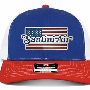 May include: A white ceramic mug with a black handle and the text "But First, Coffee" in a bold, black font. The mug is set against a blurred background, suggesting a cozy setting.