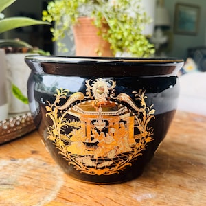 May include: A black ceramic planter with a gold decorative design. The planter features a detailed illustration of a classical scene with figures and architecture. The rim and accents are also gold.