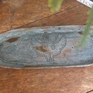 May include: A handmade, oval-shaped ceramic tray with a textured, mottled blue-green glaze. The tray features an embossed bird design in the center. The tray is displayed on a wooden surface.