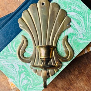 May include: A polished brass wall sconce featuring a scalloped shell-shaped backplate with a central candle holder cup and a small circular mounting hole at the top. The metal surface has a reflective gold-toned finish with curved decorative accents.