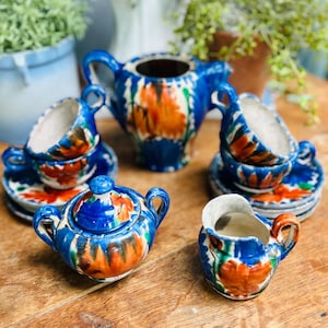 May include: A vintage ceramic tea set with a blue, orange, and white floral design. The set includes a teapot, sugar bowl, creamer, and two teacups with saucers. The tea set is displayed on a wooden surface.