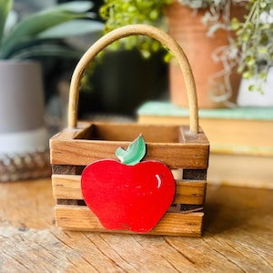May include: A small, wooden basket with a curved handle. The basket features a bright red apple with a green leaf attached to the front. The basket is made of wooden slats and is a decorative item.