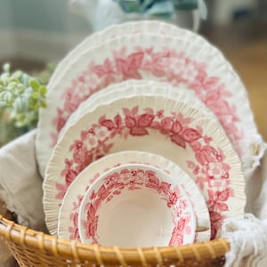 May include: A set of four vintage teacups and saucers with a pink floral pattern. The cups and saucers are stacked inside a wicker basket.