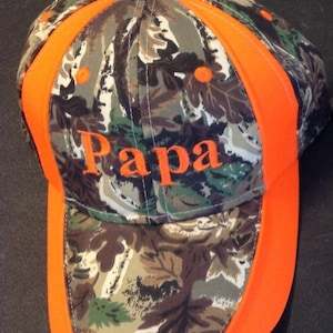 May include: Camouflage baseball cap with orange accents and the word "Papa" embroidered in orange thread. The cap features a classic six-panel design with a curved brim and an adjustable strap for a comfortable fit. The cap is made of durable fabric.
