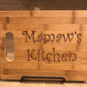 May include: A natural wood cutting board with a handle. The board is engraved with the text "Mamaw's Kitchen" in a decorative font.