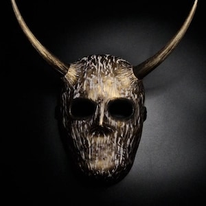 Può includere: Una maschera dettagliata con un volto a forma di teschio e lunghe corna ricurve. La maschera ha un aspetto strutturato e invecchiato con toni oro e marrone. Lo sfondo è nero, evidenziando l'intricato design della maschera.