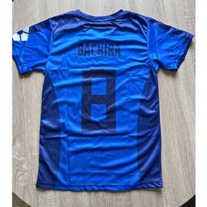 May include: Blue soccer jersey with the name "BACHIRA" and the number "8" printed on the back. The jersey has a two-tone design with darker blue accents and a white and blue logo on the sleeve. The shirt is made of a lightweight, possibly athletic material.
