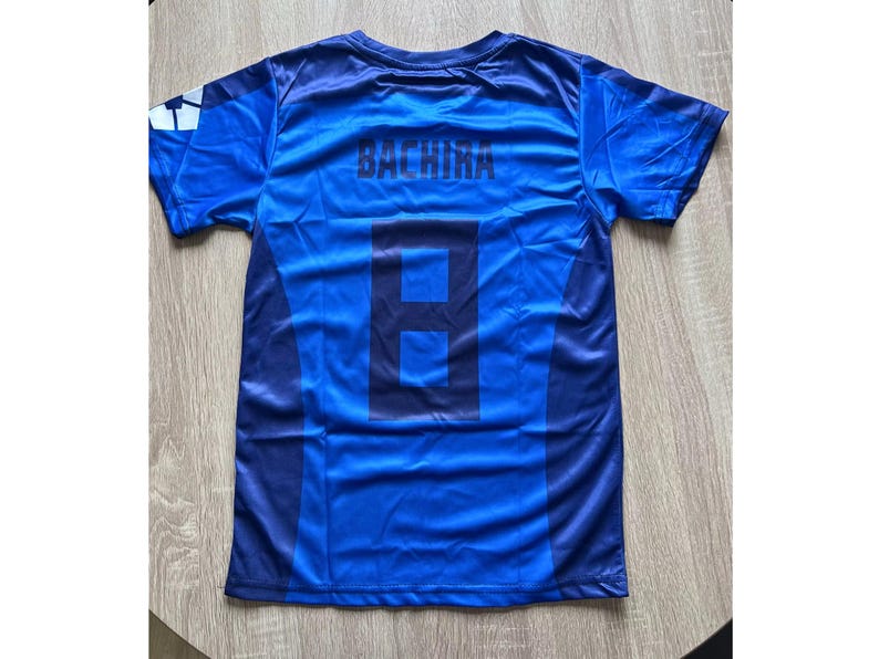 May include: Blue soccer jersey with the name "BACHIRA" and the number "8" printed on the back. The jersey has a two-tone design with darker blue accents and a white and blue logo on the sleeve. The shirt is made of a lightweight, possibly athletic material.