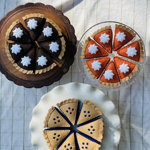 May include: Three felt pie sets arranged on serving stands. One dark brown pie with white whipped cream dollops, one orange pie with white whipped cream dollops, and one tan pie with blue filling and diamond-shaped cutouts in the crust.