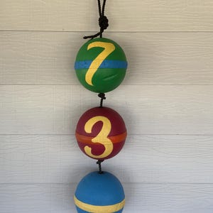 May include: Three colorful buoys hanging from a black rope. The top buoy is green with a yellow "7" and a blue stripe. The middle buoy is red with a yellow "3" and an orange stripe. The bottom buoy is blue with a yellow stripe.