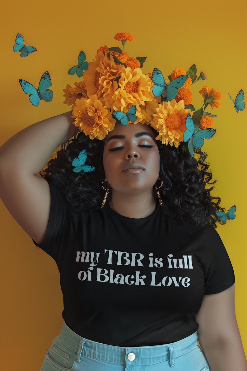 May include: A woman wearing a black t-shirt with the text "my TBR is full of Black Love." She is adorned with a floral crown of yellow flowers and orange accents, and blue butterflies. The background is a solid yellow.