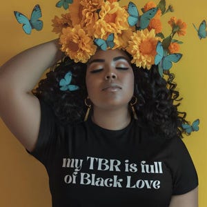 May include: A black t-shirt featuring the white text 'my TBR is full of Black Love' worn with light blue denim jeans. The person has dark curly hair adorned with large yellow flowers and several bright blue butterflies against a yellow background.