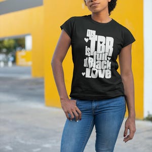 Puede incluir: Una camiseta negra de manga corta con texto blanco que dice "my TBR is full of Black Love" con pequeños iconos de corazones intercalados en las letras. La camiseta se combina con pantalones vaqueros azules y gafas de montura transparente.