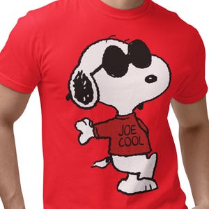 May include: A bright red short-sleeved t-shirt featuring a graphic print of the cartoon character Snoopy wearing black sunglasses and a red sweater with the text JOE COOL printed in black on the front of the sweater.