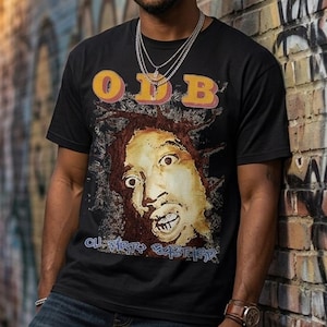 May include: Black t-shirt with a graphic of a person's face and the letters "O.D.B." in yellow and orange. The person is wearing the shirt with blue jeans and a watch. The background is a brick wall with graffiti.