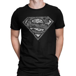 Może przedstawiać: Czarny t-shirt z postarzanym szarym logo Supermana. Kultowy symbol "S" jest widoczny na przodzie. Koszulka ma krótkie rękawy i okrągły dekolt, odpowiednia do noszenia na co dzień.