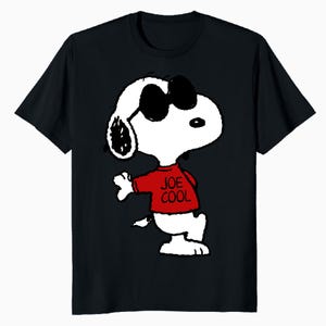May include: A black short-sleeved t-shirt featuring a graphic of the cartoon character Snoopy wearing black sunglasses and a red shirt with the text JOE COOL printed in black. The character is depicted in a standing pose with one arm extended.