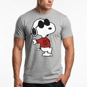 May include: A heather gray short-sleeved t-shirt featuring a graphic of the cartoon character Snoopy wearing black sunglasses and a red sweater with the text JOE COOL printed in black on the front of the sweater, centered on the chest area.