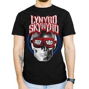 Pode incluir: Camiseta preta com um design de caveira usando óculos vermelhos, as palavras "FREE BIRD" e o nome da banda "LYNYRD SKYNYRD" em uma fonte desgastada. Um círculo azul envolve o design.