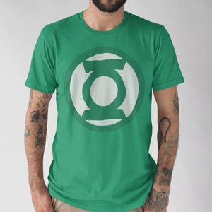 Peut inclure: T-shirt vert avec le logo blanc de Green Lantern sur le devant. Le logo est un design stylisé d'une lanterne avec un centre circulaire. Le t-shirt est de couleur verte unie et a des manches courtes.