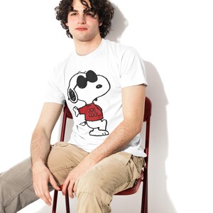 May include: A white short-sleeved t-shirt featuring a graphic print of the cartoon character Snoopy wearing black sunglasses and a red sweater with the text "JOE COOL" printed in black on the front of the sweater, paired with tan cargo pants.