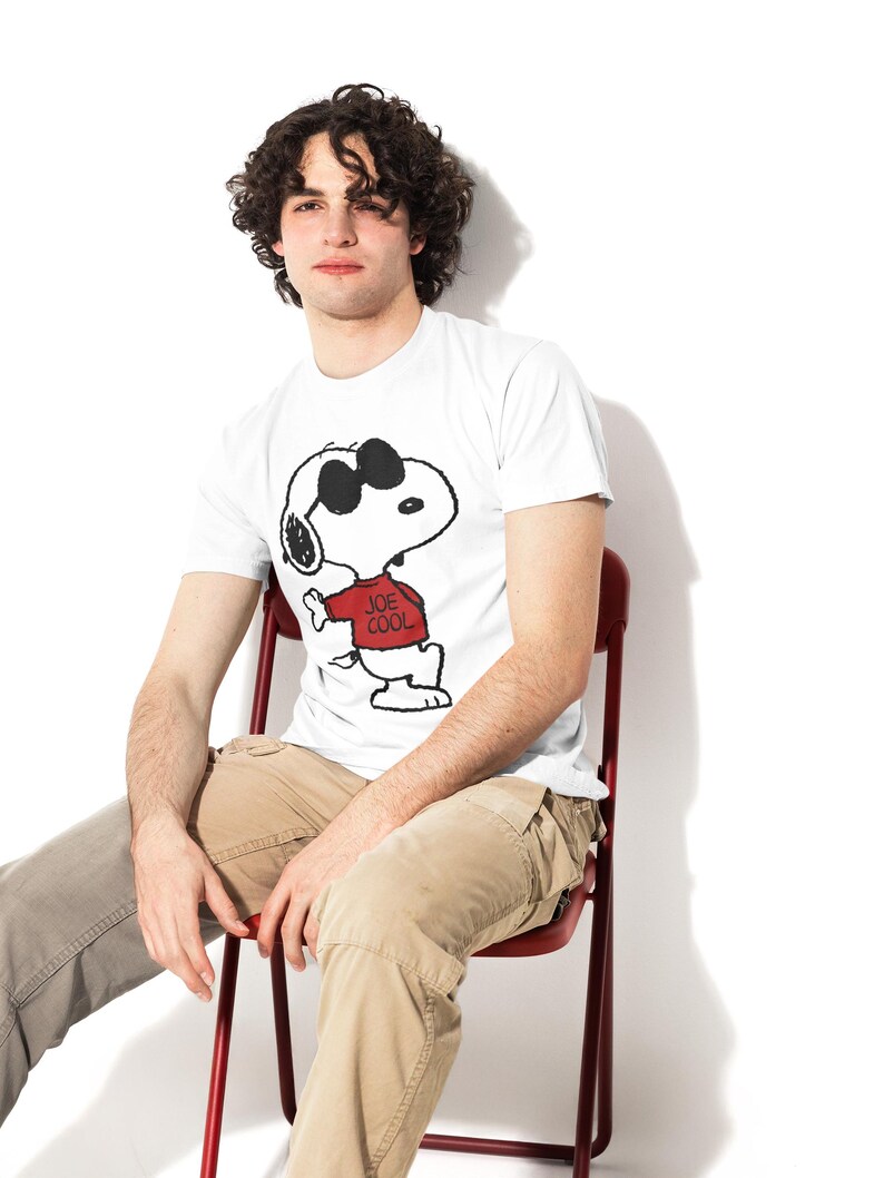 May include: A white short-sleeved t-shirt featuring a graphic print of the cartoon character Snoopy wearing black sunglasses and a red sweater with the text "JOE COOL" printed in black on the front of the sweater, paired with tan cargo pants.