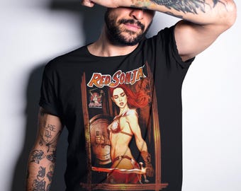 May include: Black t-shirt featuring a graphic print of Red Sonja, a fictional sword-wielding warrior, holding a sword and shield. The graphic is in a comic book style with a red, orange, and yellow colour scheme.