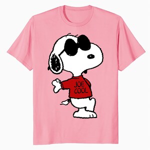 May include: A light pink short-sleeved t-shirt featuring a graphic of the cartoon character Snoopy wearing black sunglasses and a red shirt with the text JOE COOL printed in black. The character is depicted in a white and black line art style.