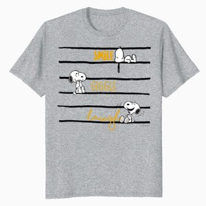 Puede incluir: Una camiseta gris con Snoopy de Peanuts. La camiseta tiene líneas horizontales negras con las palabras "Smile", "Giggle" y "Laugh" en amarillo. Snoopy se representa en varias poses encima y debajo de las líneas.