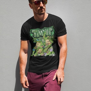 May include: A black short-sleeved t-shirt featuring a graphic print of the Teenage Mutant Ninja Turtles with the text TMNT in a green graffiti-style font. The design includes the four turtles in action poses against a green spray-painted background.