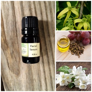 May include: A small brown glass bottle with a black cap, labeled "Facial Serum" and "0.20 oz". The bottle is sitting on a wooden surface. The image also includes close-ups of various ingredients, including ylang ylang flowers, grapes, grape seeds, and jasmine flowers.