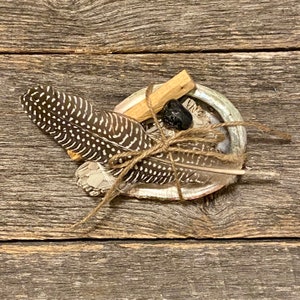May include: A white abalone shell filled with sage, palo santo wood, a black stone, and a brown and white feather, all tied together with twine.