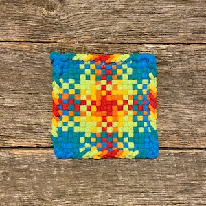 May include: A colorful woven square pot holder with a red, orange, yellow, green, and blue pattern. The pot holder is made of yarn and has a square shape.