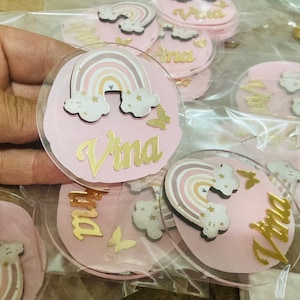Puede incluir: Un conjunto de adornos acrílicos personalizados en rosa y dorado con un diseño de arcoíris y estrellas. Cada adorno tiene un nombre diferente, incluyendo "Vina".