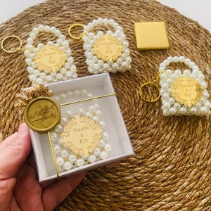 Puede incluir: Un surtido de llaveros con forma de bolso con cuentas de perlas y detalles dorados, junto con una caja de regalo. Los llaveros tienen una etiqueta dorada con los nombres "Natalia & Key". La caja de regalo está sellada con un sello de cera dorado que dice "With You".