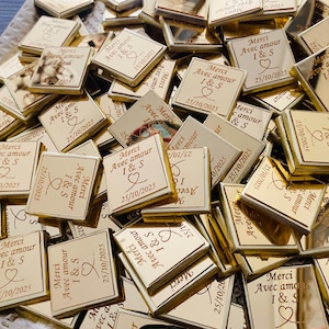 Puede incluir: Una colección de recuerdos cuadrados de color dorado. Cada recuerdo está grabado con las palabras "Merci Avec amour I & S" y la fecha "25/10/2025" junto con un símbolo de corazón. Los recuerdos están esparcidos sobre una superficie reflectante.