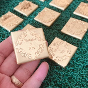 Puede incluir: Favores de chocolate de color dorado con un diseño intrincado y el texto "Natalia & Key" en la parte superior.